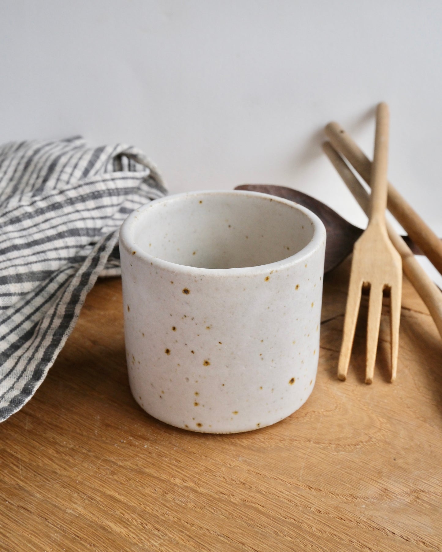 Speckled ceramic mug on a wooden surface with a striped towel and wooden utensils in the background.
