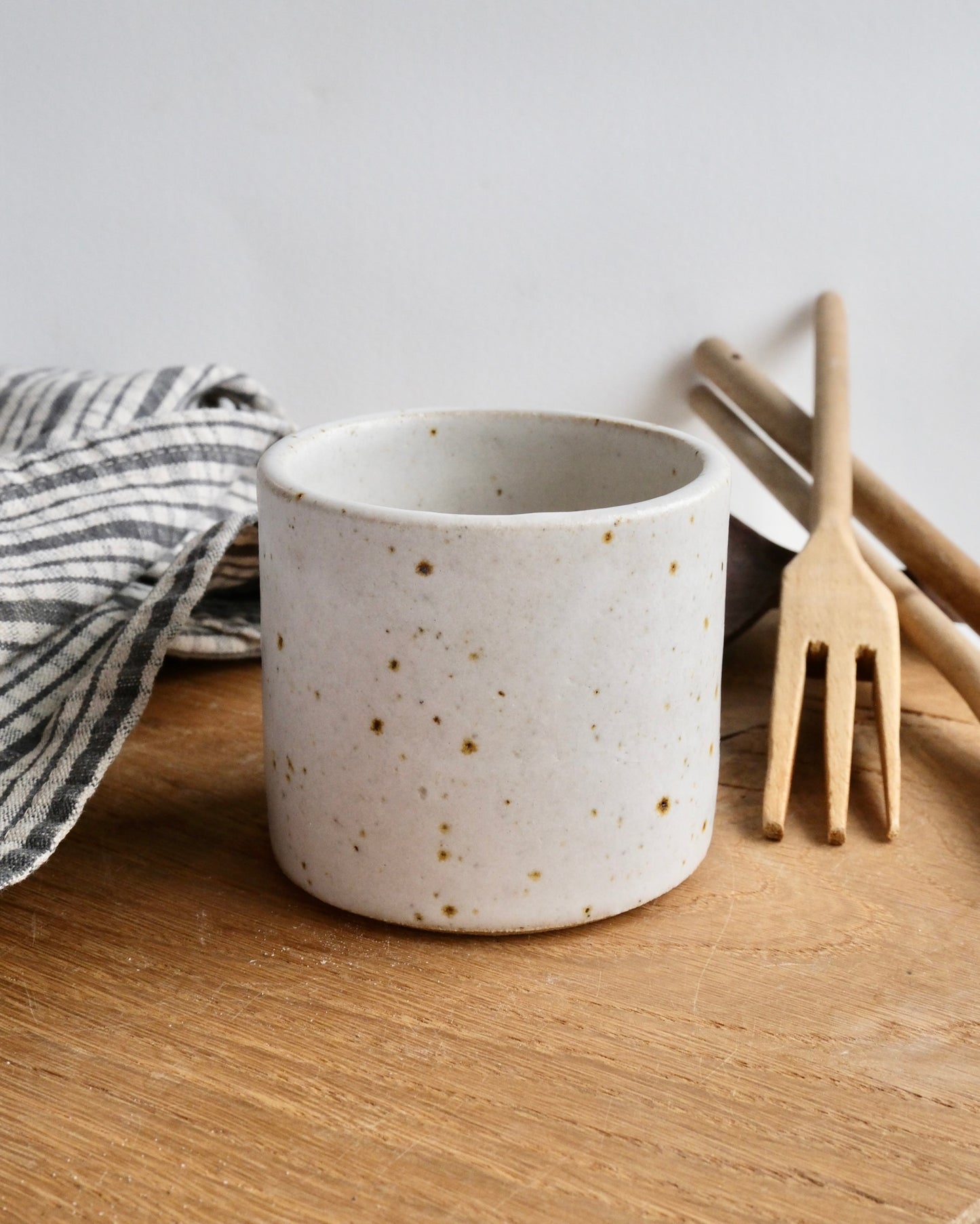 Speckled ceramic mug on a wooden surface with a striped towel and wooden utensils in the background.