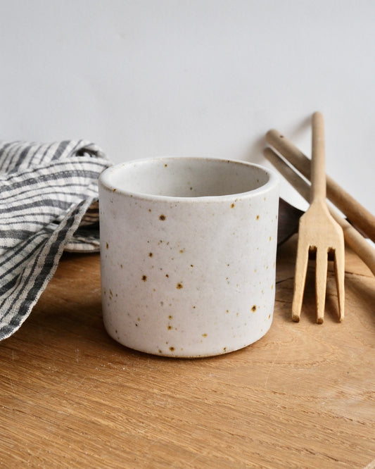 Speckled ceramic mug on a wooden surface with a striped towel and wooden utensils in the background.