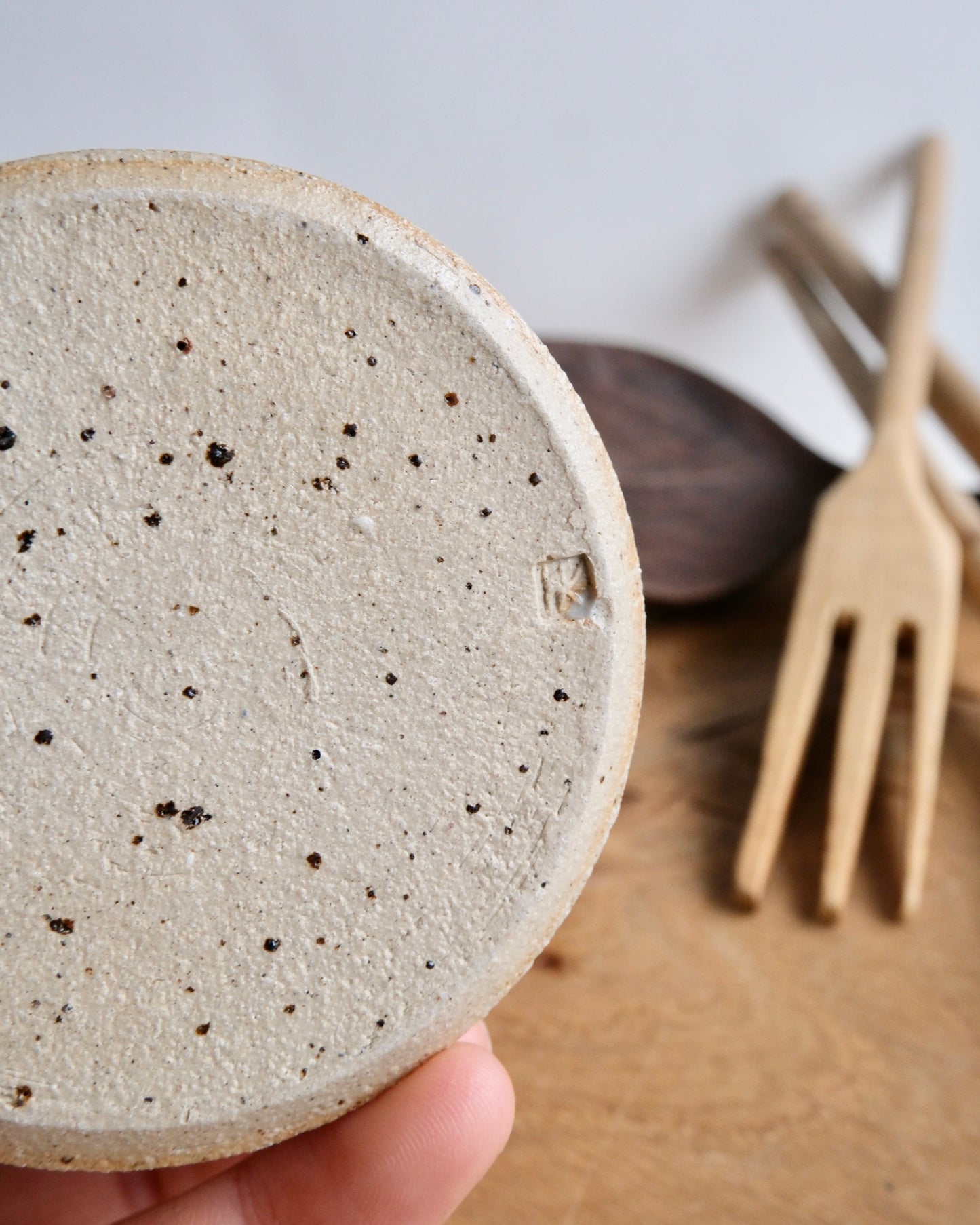 Close-up of a textured ceramic coaster held by a hand, with a blurred background featuring a wooden spoon and fork.