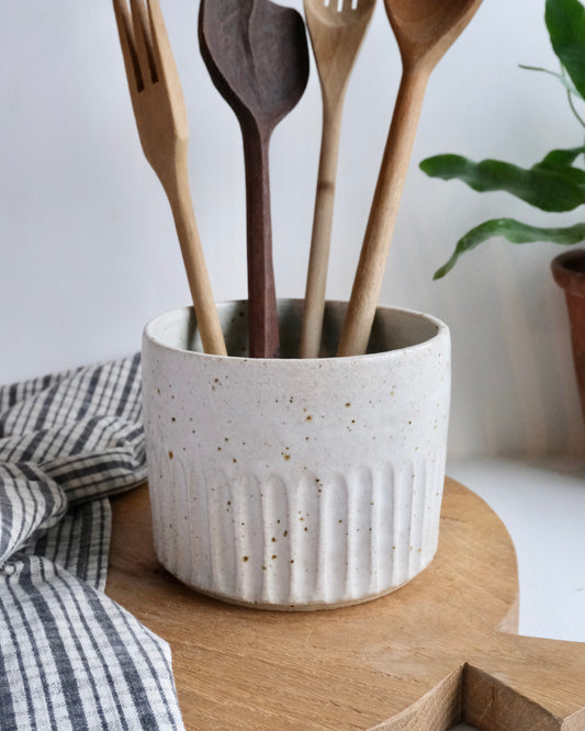 Handmade White Ceramic utensil pot with a carved line design on a wooden chopping board with wooden spoons and a tea towel. 