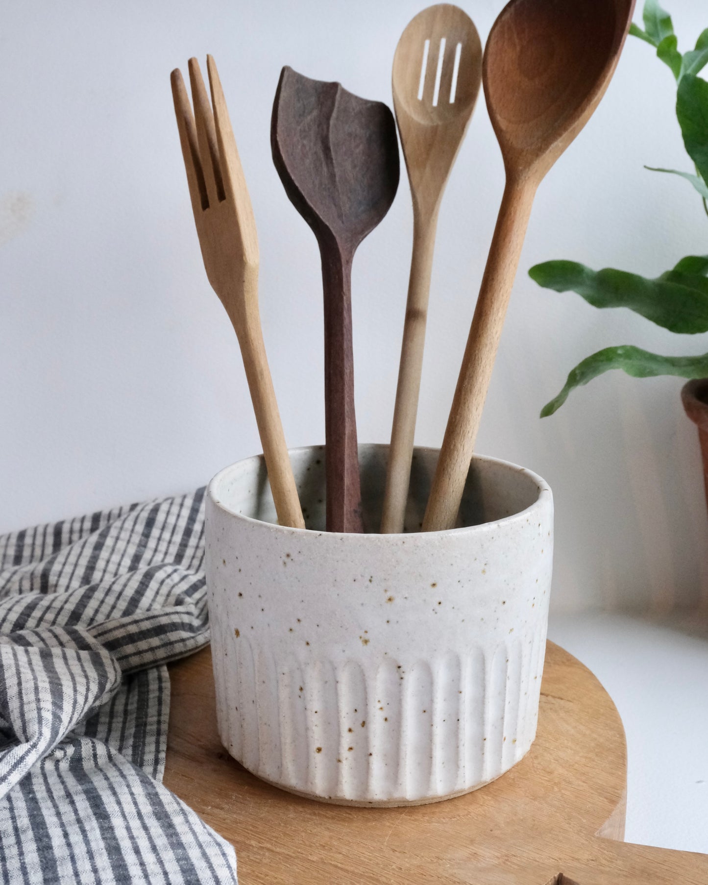 A white ceramic pot with a carved design with wooden spoons inside on a wooden chopping board and a tea towel in the background