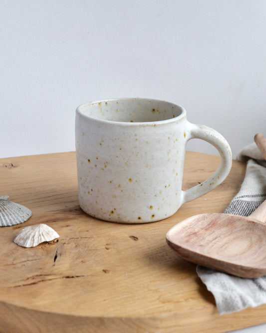A whit speckled handmade mug with a straight and simple form on a chopping board with wooden spoons.