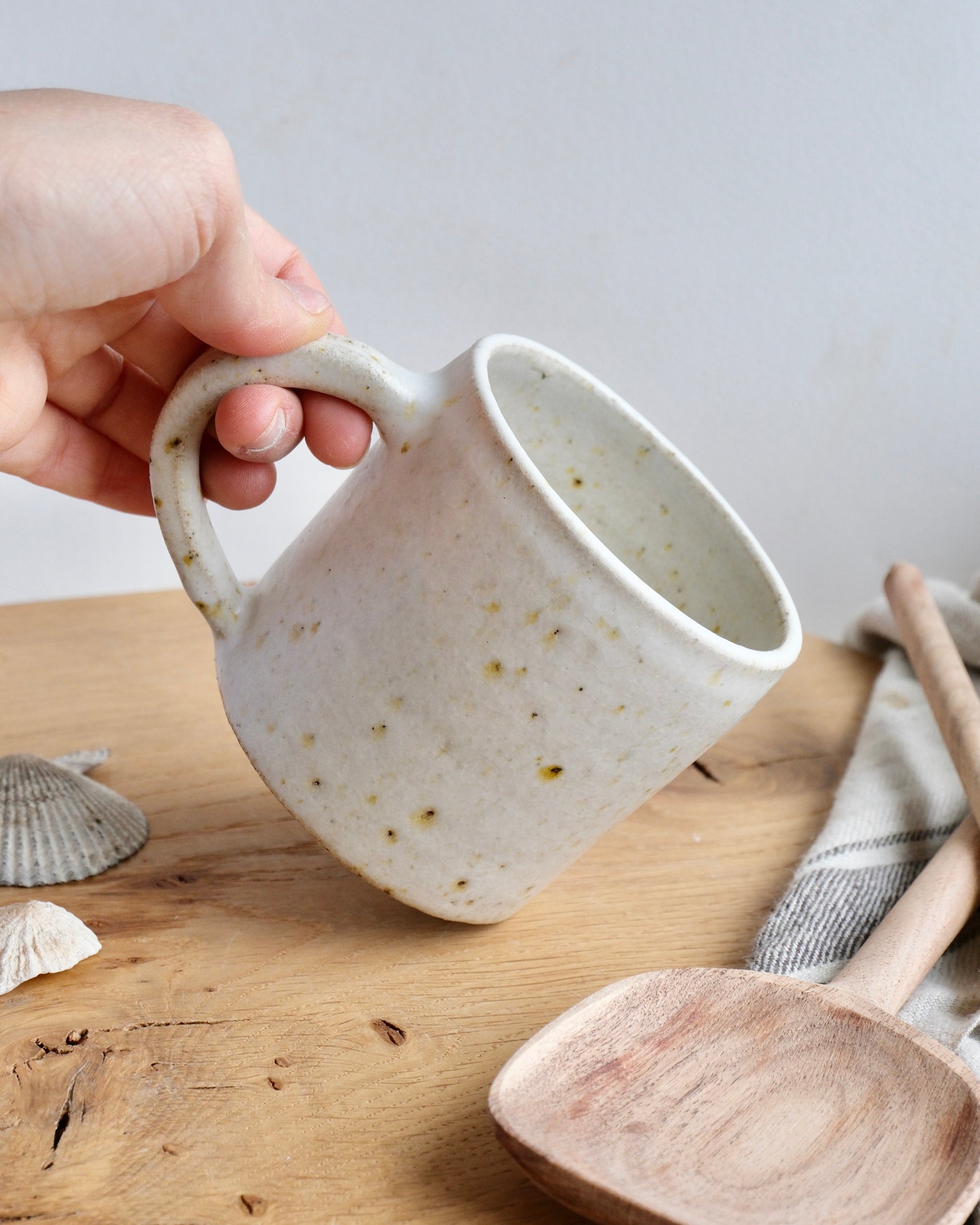 Hand holding a white ceramic mug with speckled texture glaze on a wooden surface.