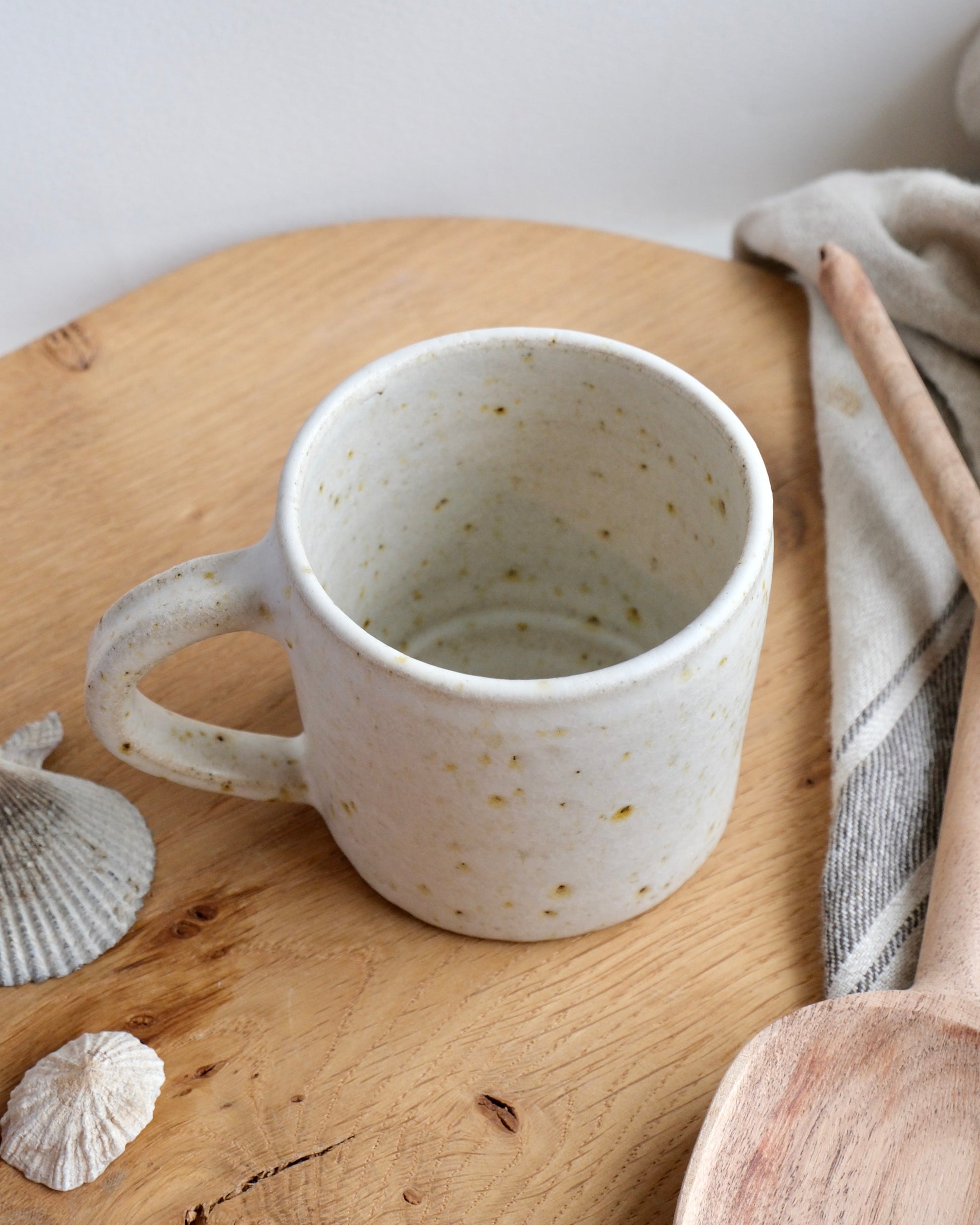 White ceramic mug on a wooden surface with a wooden spoon and shells.