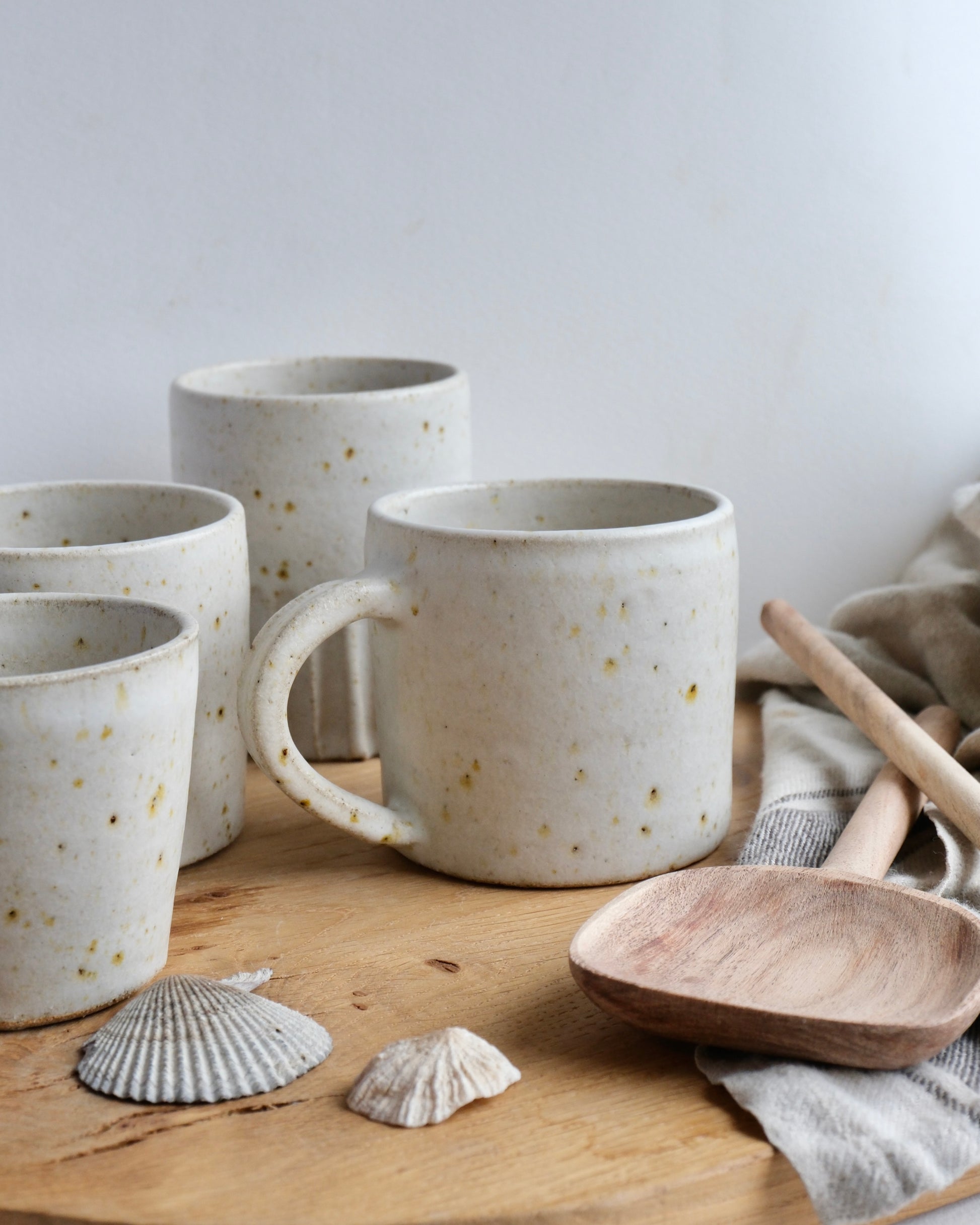 Set of white ceramic tea/ coffee mugs with speckled pattern on a wooden surface with a wooden spoon and shells.
