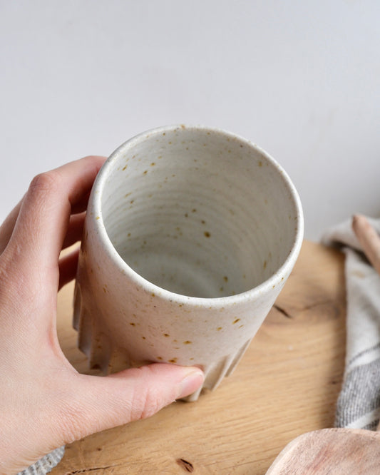 hand holding a white ceramic cup with textured design on a wooden surface with shells and a wooden spoon.