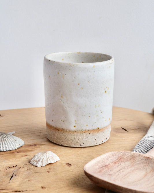 A ceramic white speckled tall toothbrush pot on a wooden chopping board 