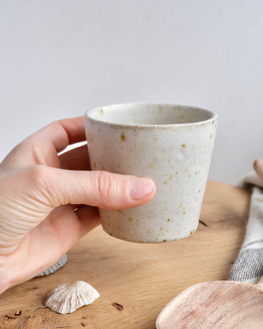 A hand holding a white speckled stoneware cup on a wooden chopping board 