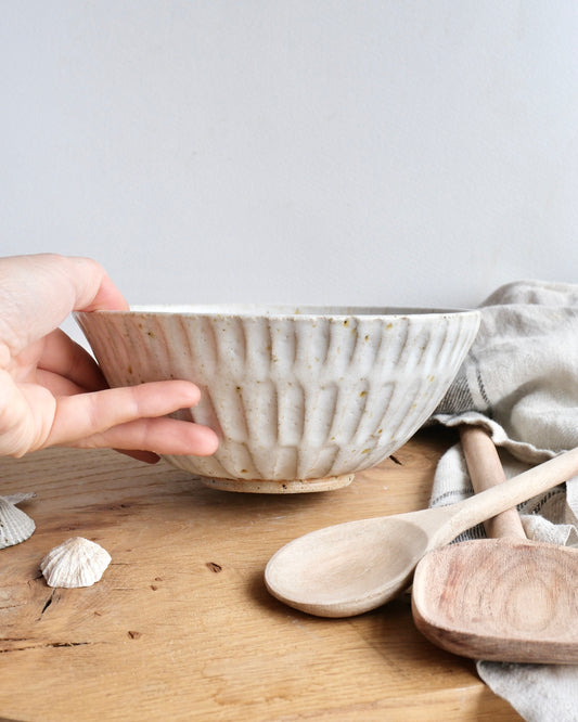 Hand holding a handmade white stoneware breakfast bowl with a textured carved design on a wooden chopping board
