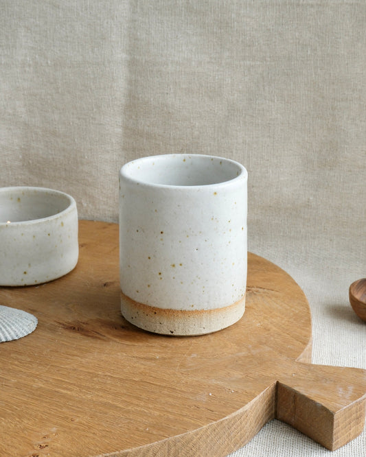 A handmade ceramic pencil pot with a speckled off-white glaze and a natural tan base, displayed on a wooden board against a linen background.