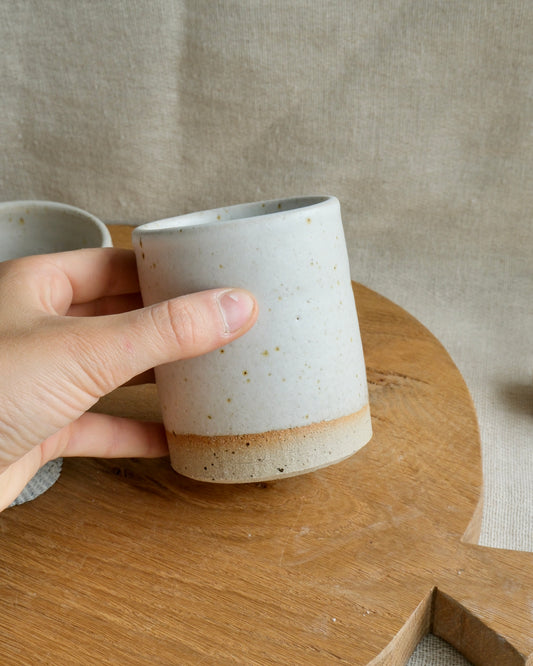 A hand holds a small handmade ceramic toothbrush holder with a speckled off-white glaze and a natural tan unglazed band at the base, set on a wooden board against a linen backdrop.