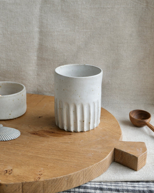 A speckled off-white ceramic cup with a carved ribbed base, displayed on a round wooden serving board against a neutral linen background.