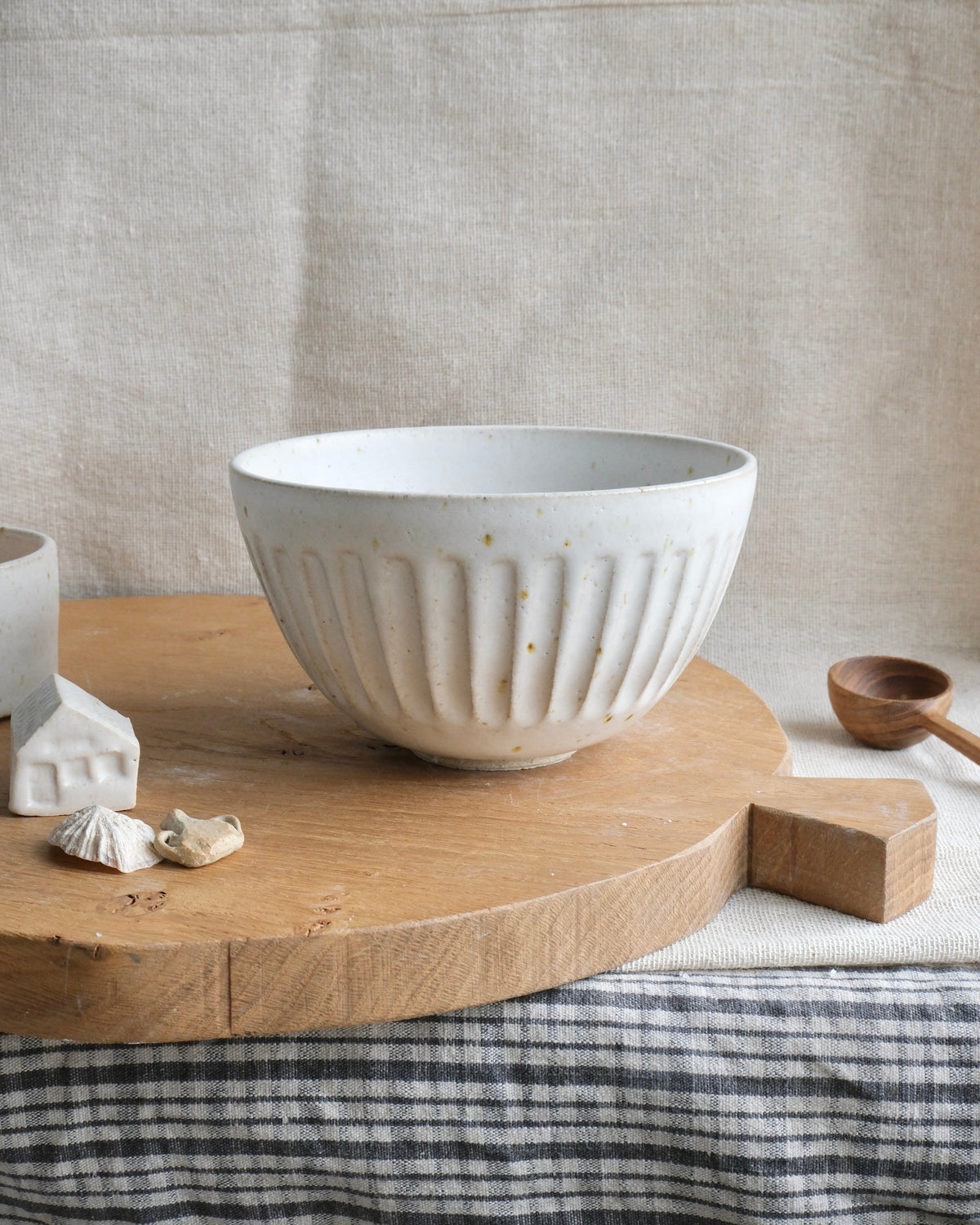 A handmade ceramic bowl with a ribbed fluted exterior and speckled off-white glaze, displayed on a round wooden serving board with a striped cloth underneath.