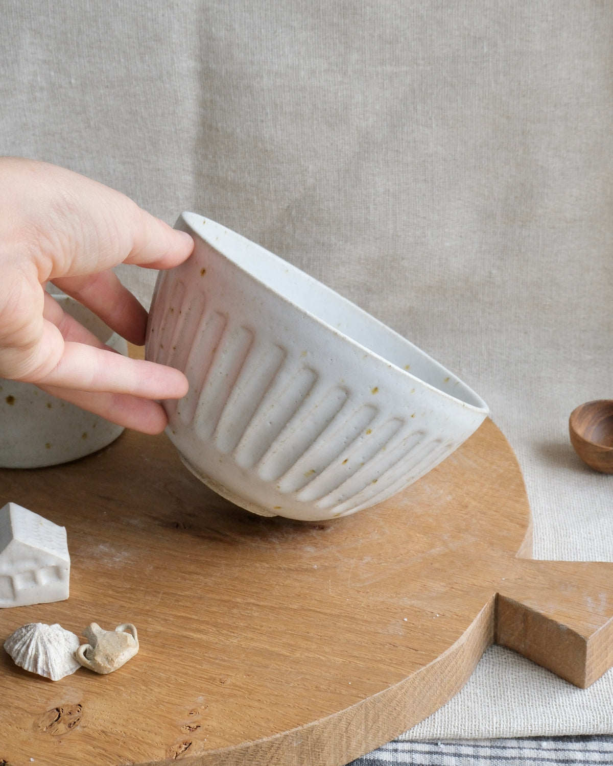 A hand holds a small ribbed ceramic cereal bowl with speckled beige and white glaze, set on a round wooden board against a neutral linen backdrop.