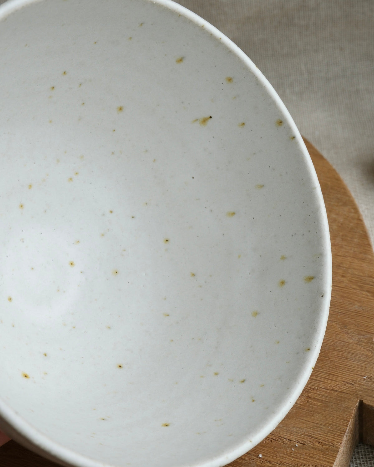 A close-up of a white ceramic bowl with small brown speckles, shown resting on a wooden board against a neutral fabric background.