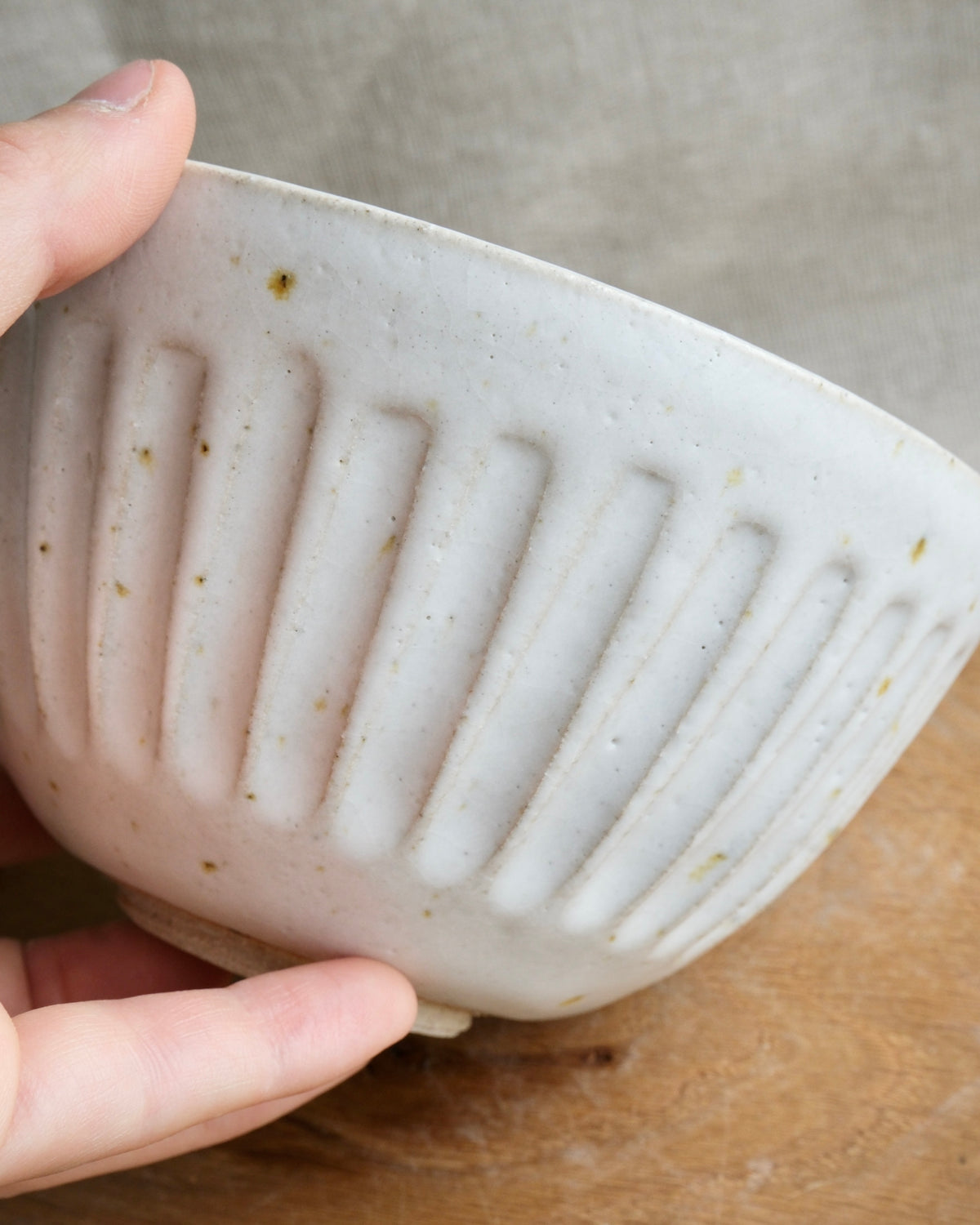 A close-up of a hand holding a speckled off-white ceramic bowl with vertical ribbed detailing, set against a neutral gray background and a wooden surface.