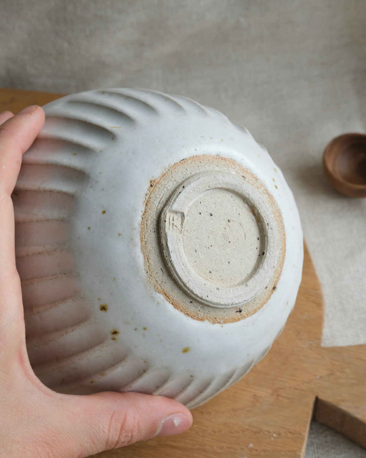 A hand holds a handmade ceramic bowl upside down to show the textured, ribbed bottom and speckled glaze. The bowl has an off-white glaze with a natural beige clay base ring.