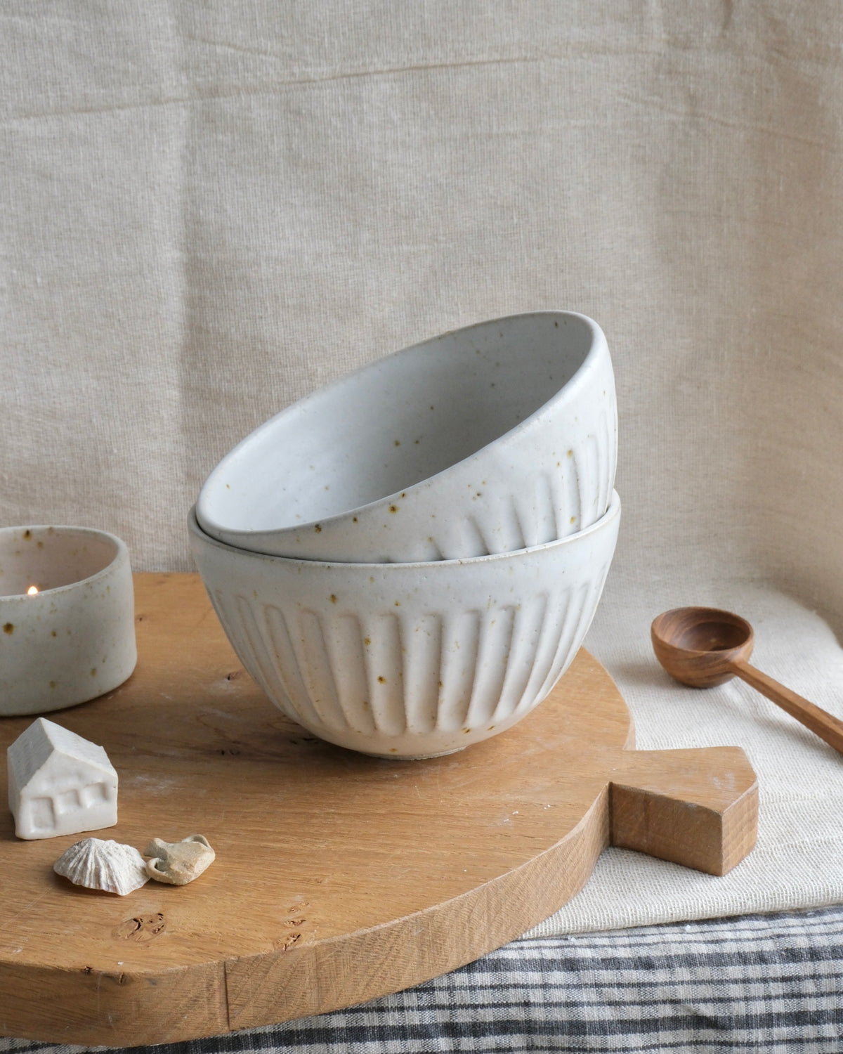 two stacked ceramic bowls with a ribbed exterior and speckled cream glaze, displayed on a round wooden board with small decorative props against a linen backdrop.