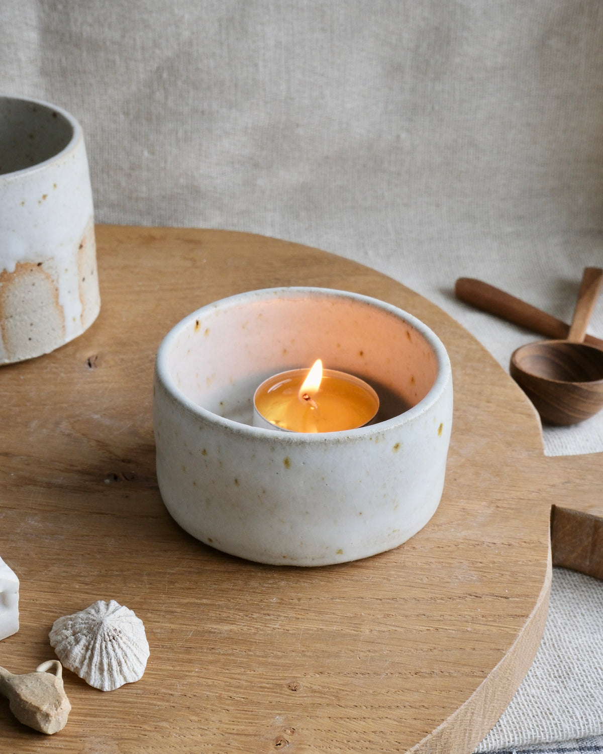 A small lit tealight candle sits inside a round, speckled off-white ceramic holder on a wooden board, with rustic neutral props in the background.