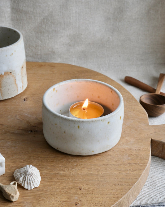 A small lit tealight candle sits inside a round, speckled off-white ceramic holder on a wooden board, with rustic neutral props in the background.