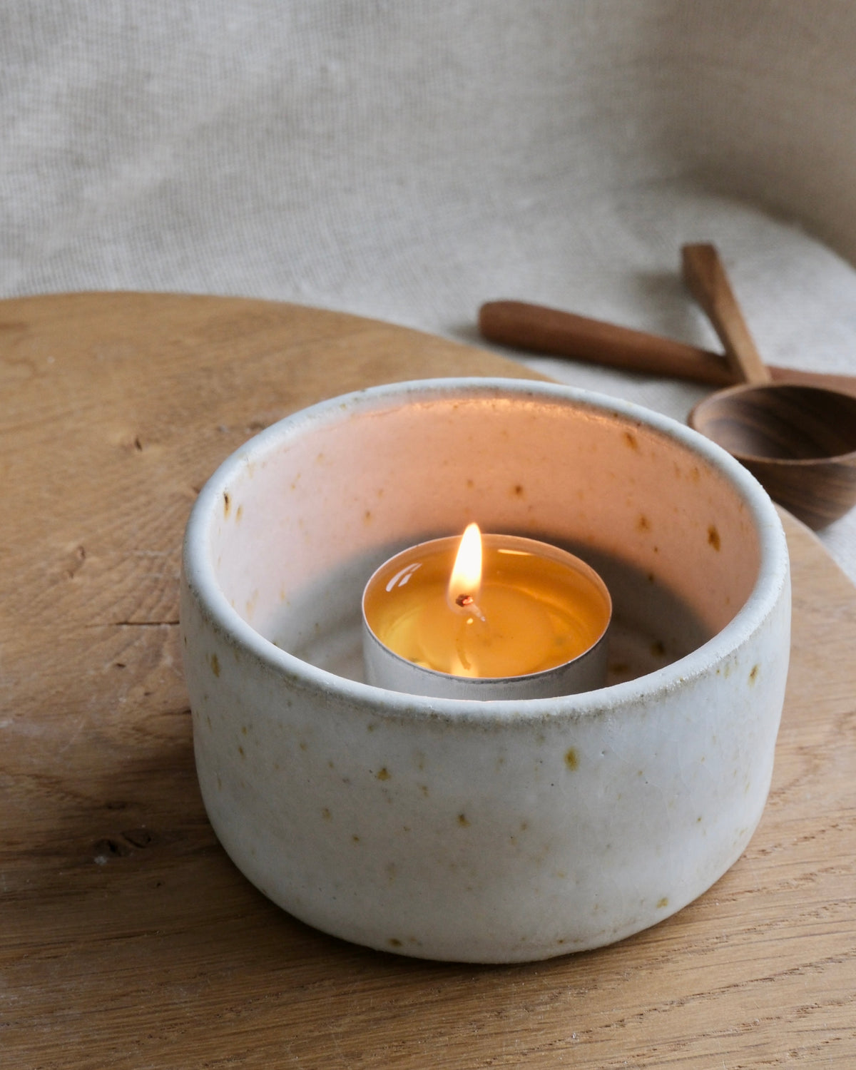 A lit tea light candle sits inside a round, speckled off-white ceramic holder on a wooden surface, with a softly blurred neutral background and small wooden props.