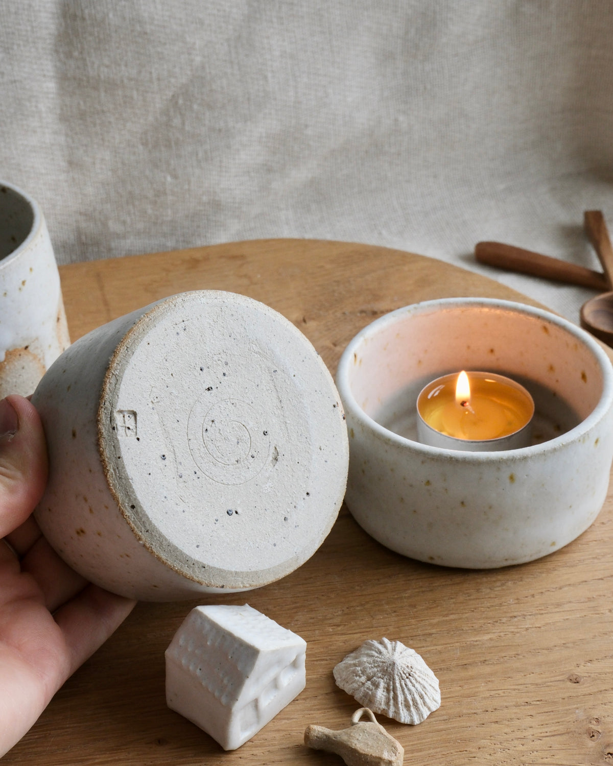 A hand holds a speckled off-white ceramic candle holder over a wooden table, next to another matching holder containing a lit tealight candle. The scene includes small decorative objects and a linen backdrop.