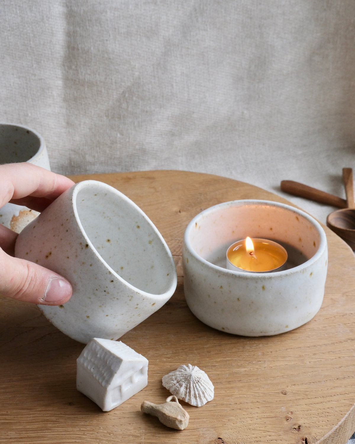A hand holds a speckled off-white ceramic dish over a wooden table, next to a matching ceramic candle holder with a lit tea light candle. Small decorative objects, including a miniature house and a seashell, sit in front on the table against a neutral linen backdrop.