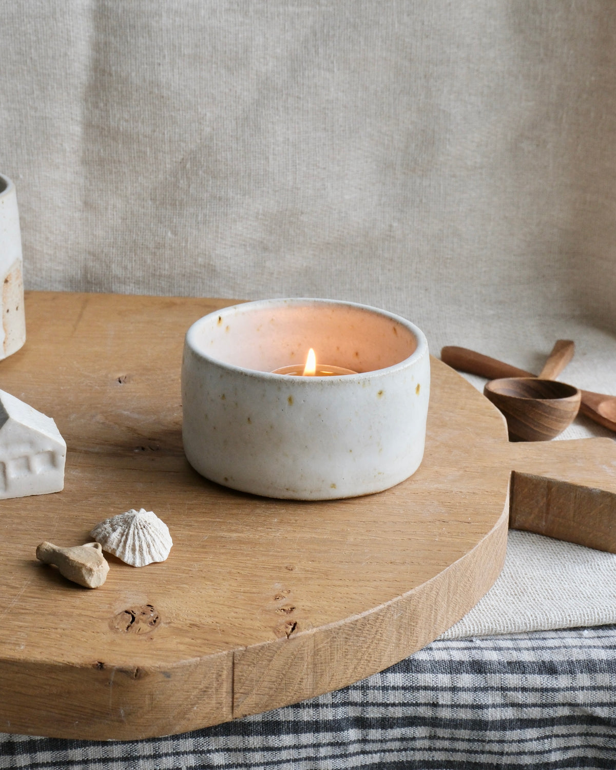 A small white ceramic candle holder with a lit candle, styled on a round wooden board with linen fabric and rustic props in the background.