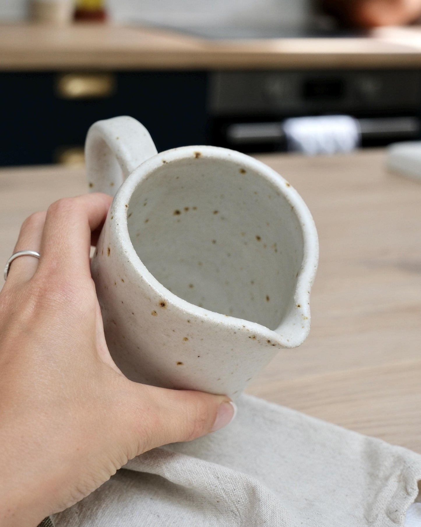 Hand holding a medium white speckled ceramic milk jug creamer with a textured surface on a wooden table.