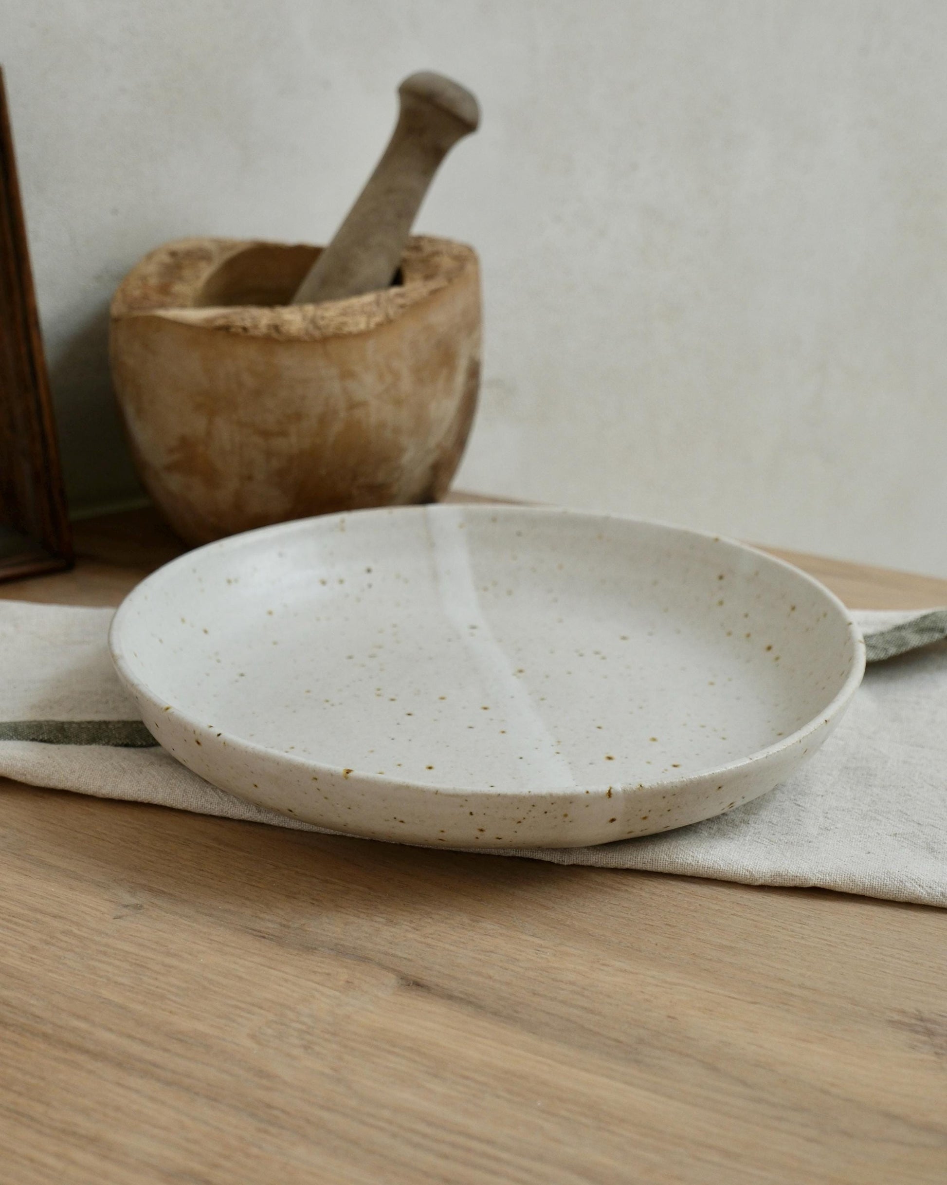 White Ceramic pasta bowl with speckled pattern on a wooden surface with a wooden mortar and pestle in the background.