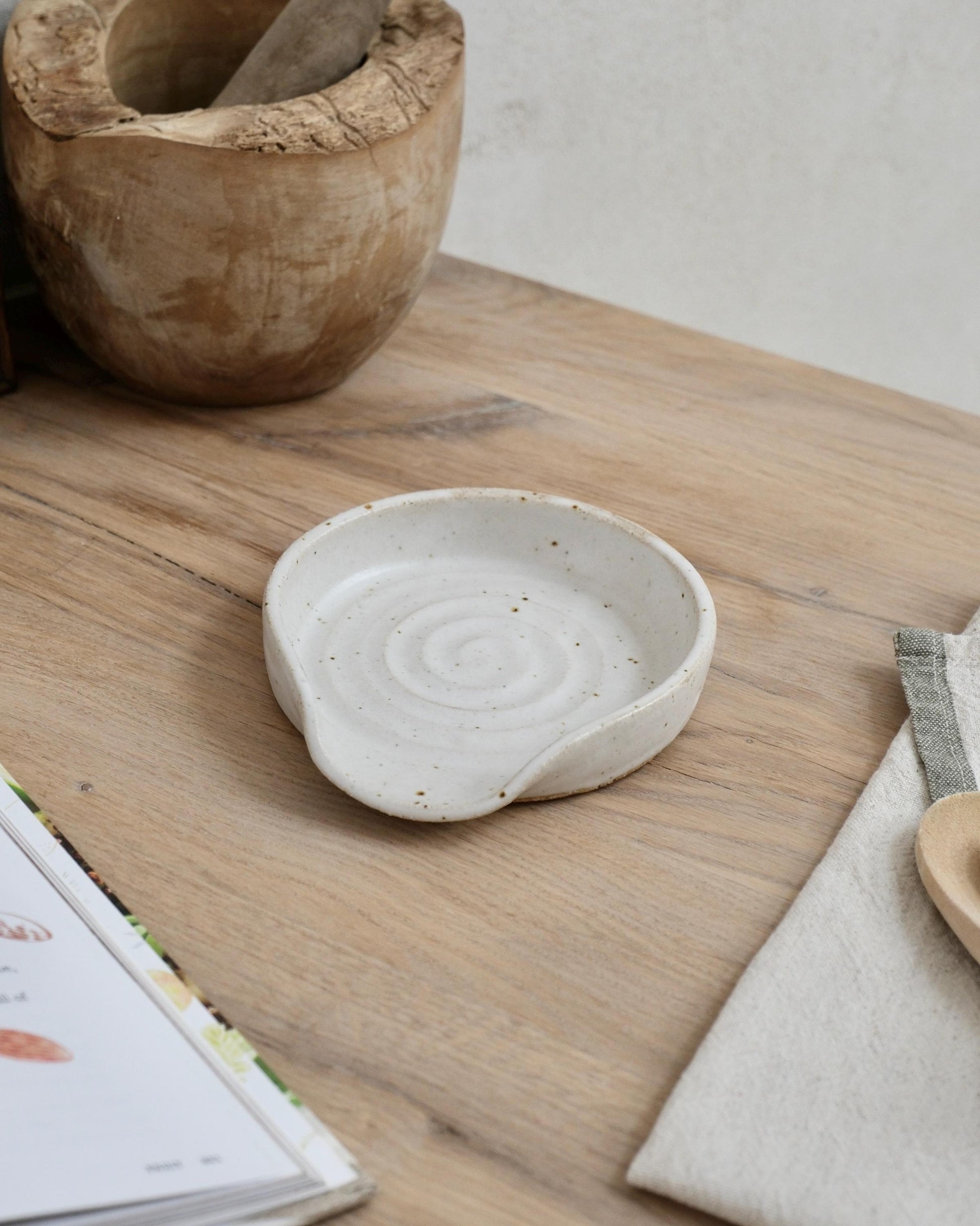 white Ceramic spoon rest dish on a wooden surface with a wooden container and book in the background