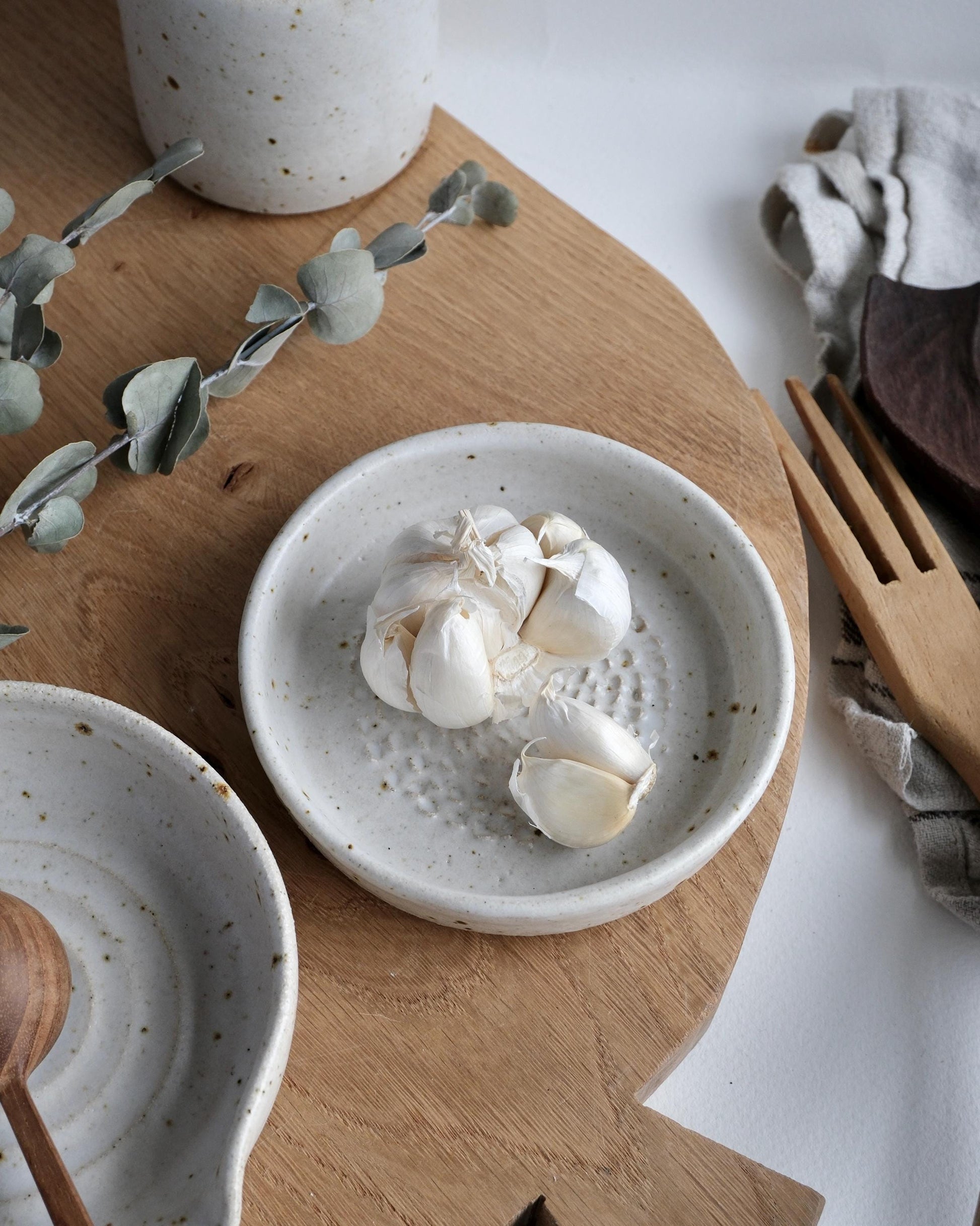 White ceramic grater dish with garlic cloves on a wooden surface with eucalyptus leaves.