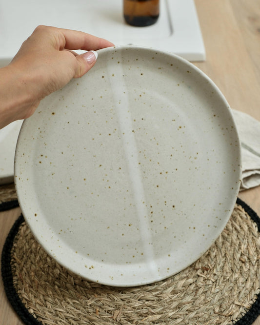 Hand holding a handmade white ceramic plate with a textured surface on a woven mat.
