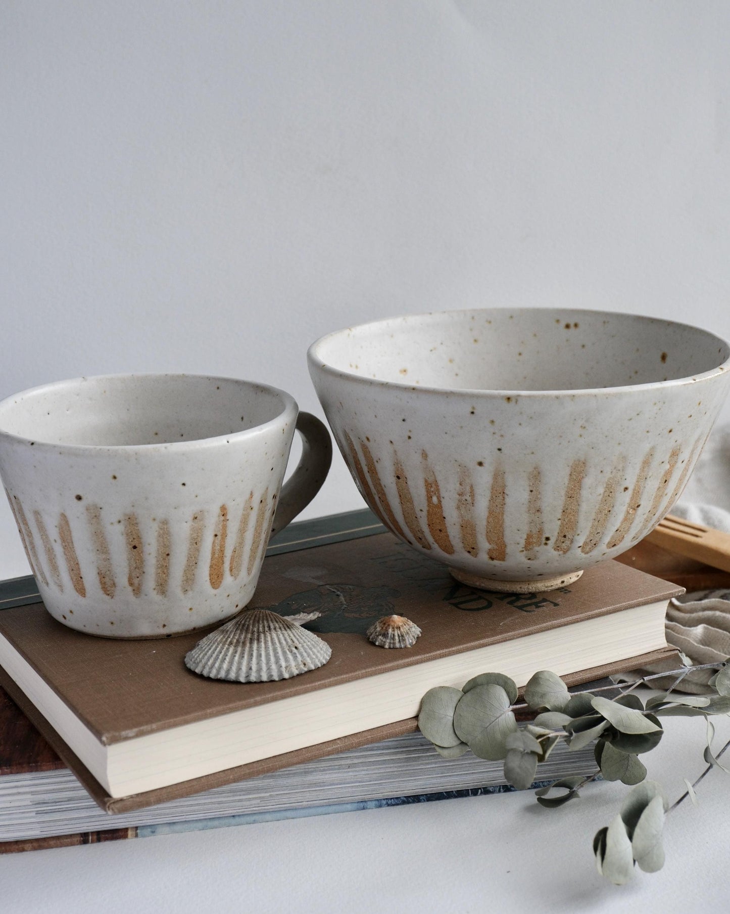 A ceramic bowl and mug with an orange striped pattern with a textured finish on a stack of books with seashells and eucalyptus leaves.