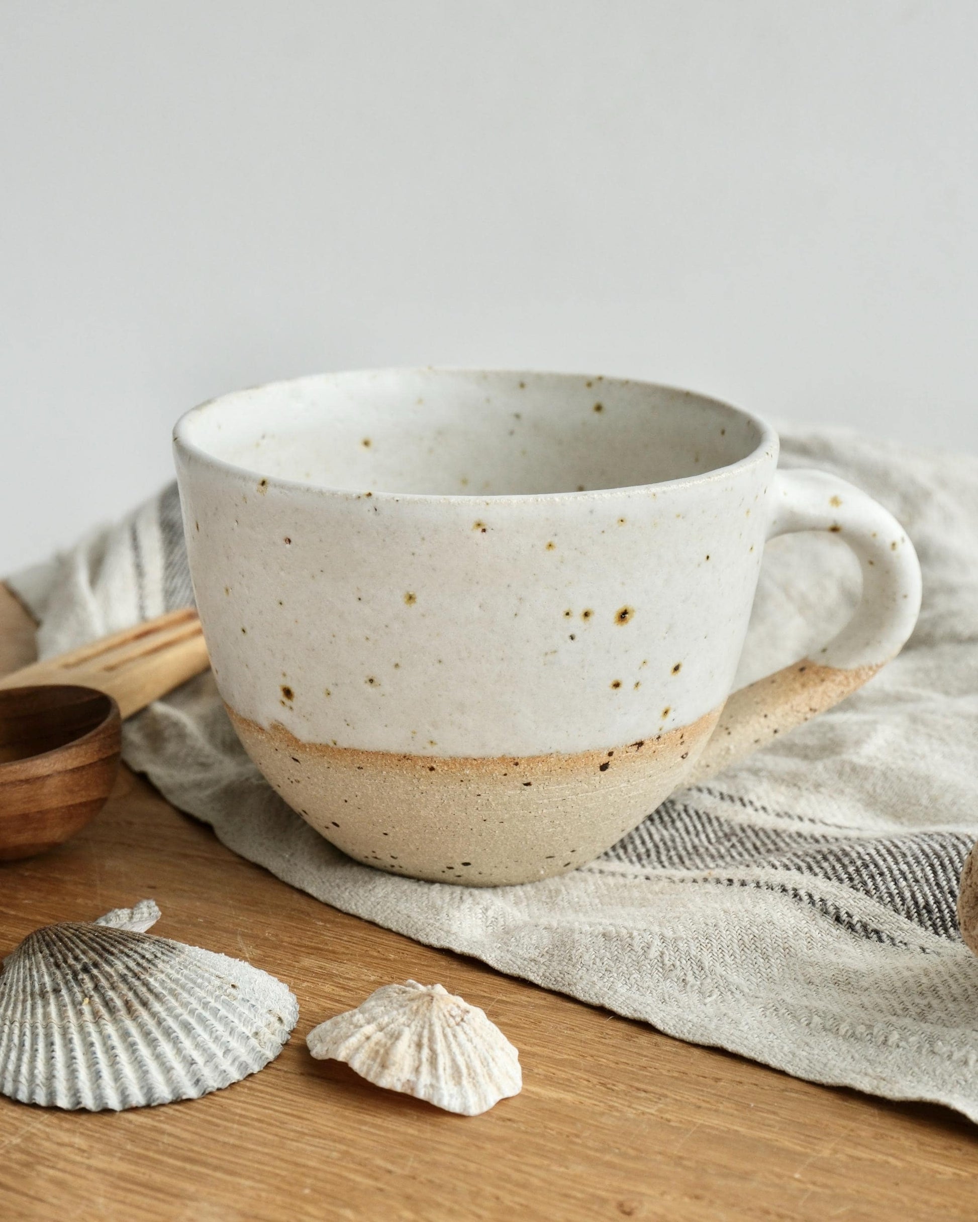white Speckled ceramic mug on a wooden surface with shells and a towel.