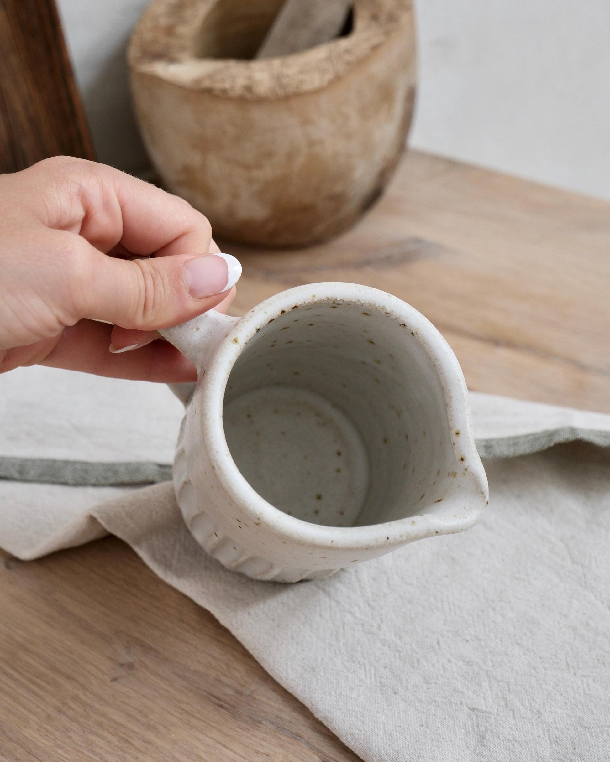 Hand holding a small ceramic pitcher milk jug on a wooden surface with a wooden bowl in the background.