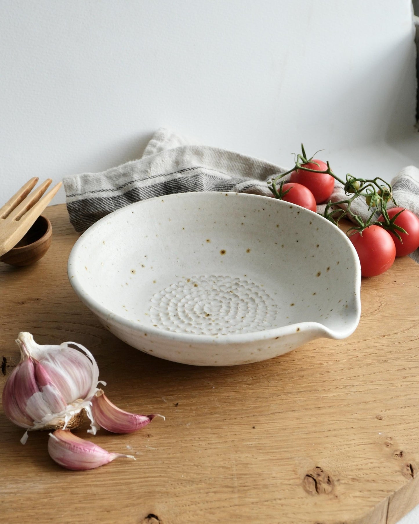 White ceramic grater bowl on a wooden surface with garlic, tomatoes, and a towel in the background.