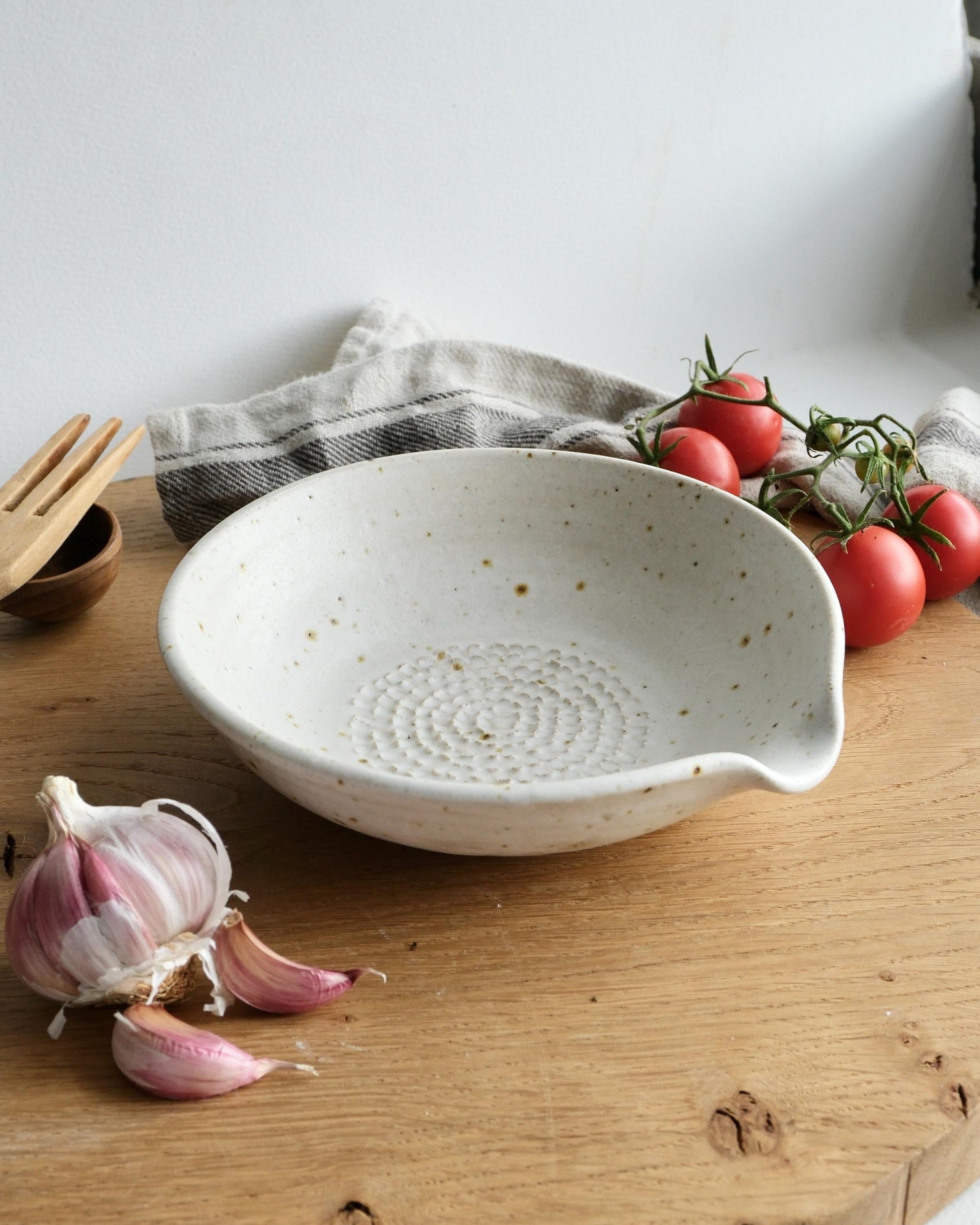 White ceramic grater bowl on a wooden surface with garlic, tomatoes, and a towel in the background.