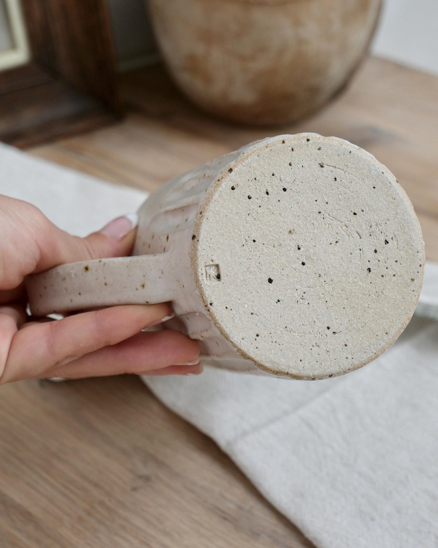 Hand holding a white speckled ceramic milk jug with a textured surface on a wooden table.
