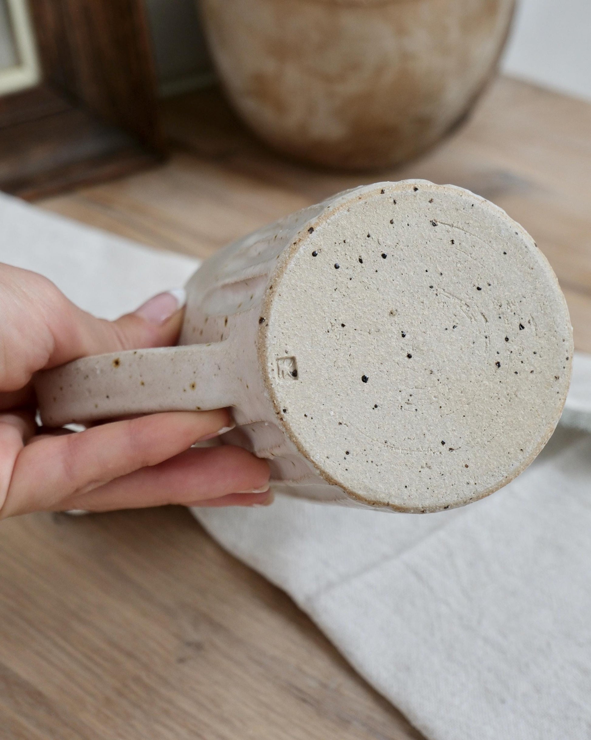 Hand holding a white speckled ceramic milk jug with a textured surface on a wooden table.