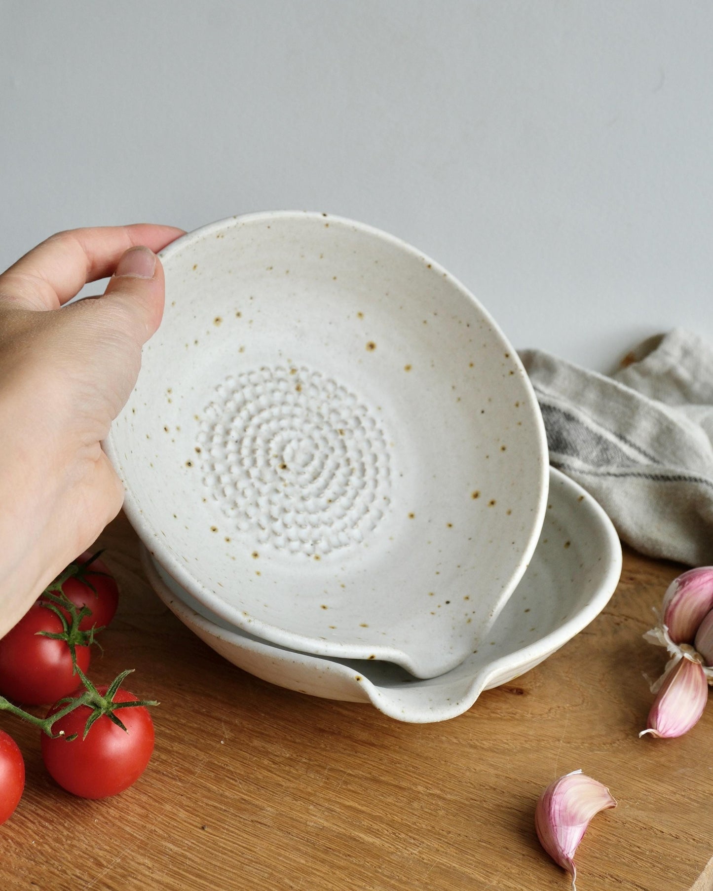 White ceramic bowl with textured grater surface held by a hand on a wooden surface with tomatoes and garlic.