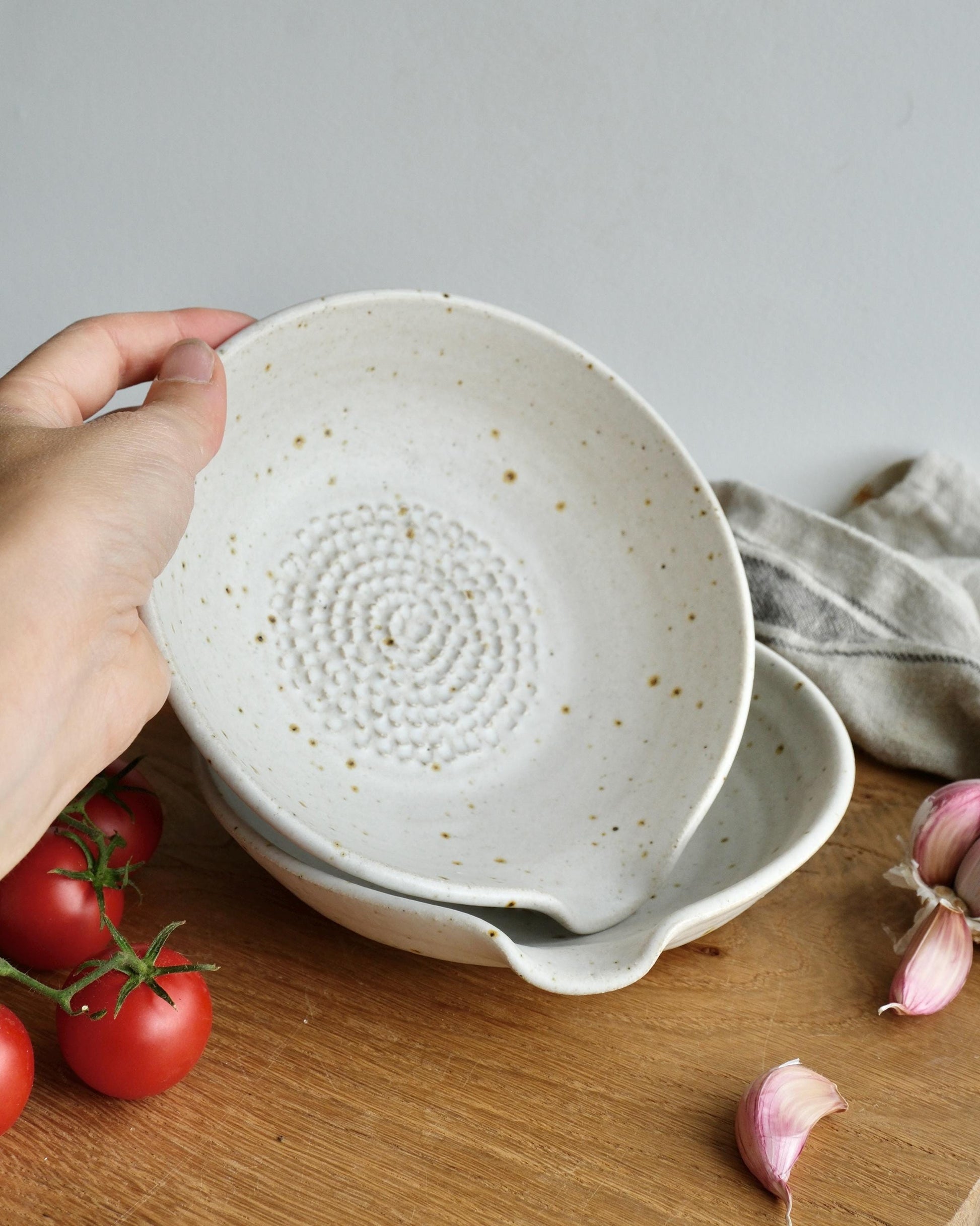 White ceramic bowl with textured grater surface held by a hand on a wooden surface with tomatoes and garlic.