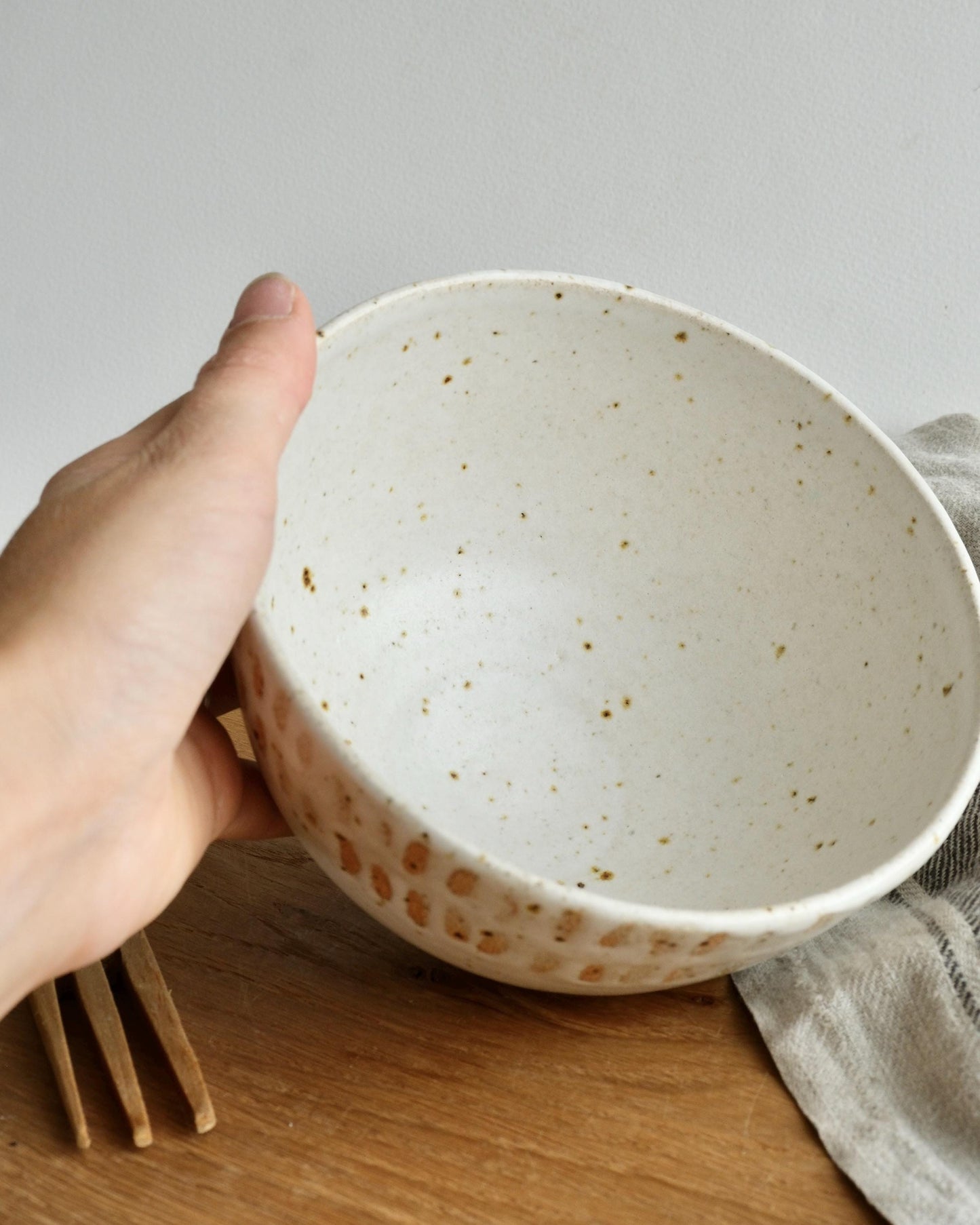 Hand holding a white speckled ceramic breakfast bowl on a wooden surface with a neutral background