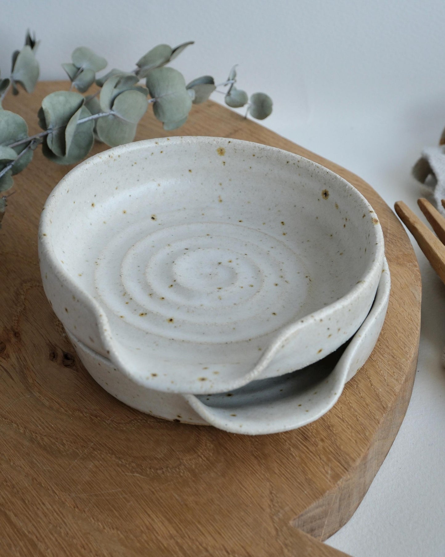 a pair of White ceramic spoon rests with speckled texture on a wooden surface with eucalyptus leaves.