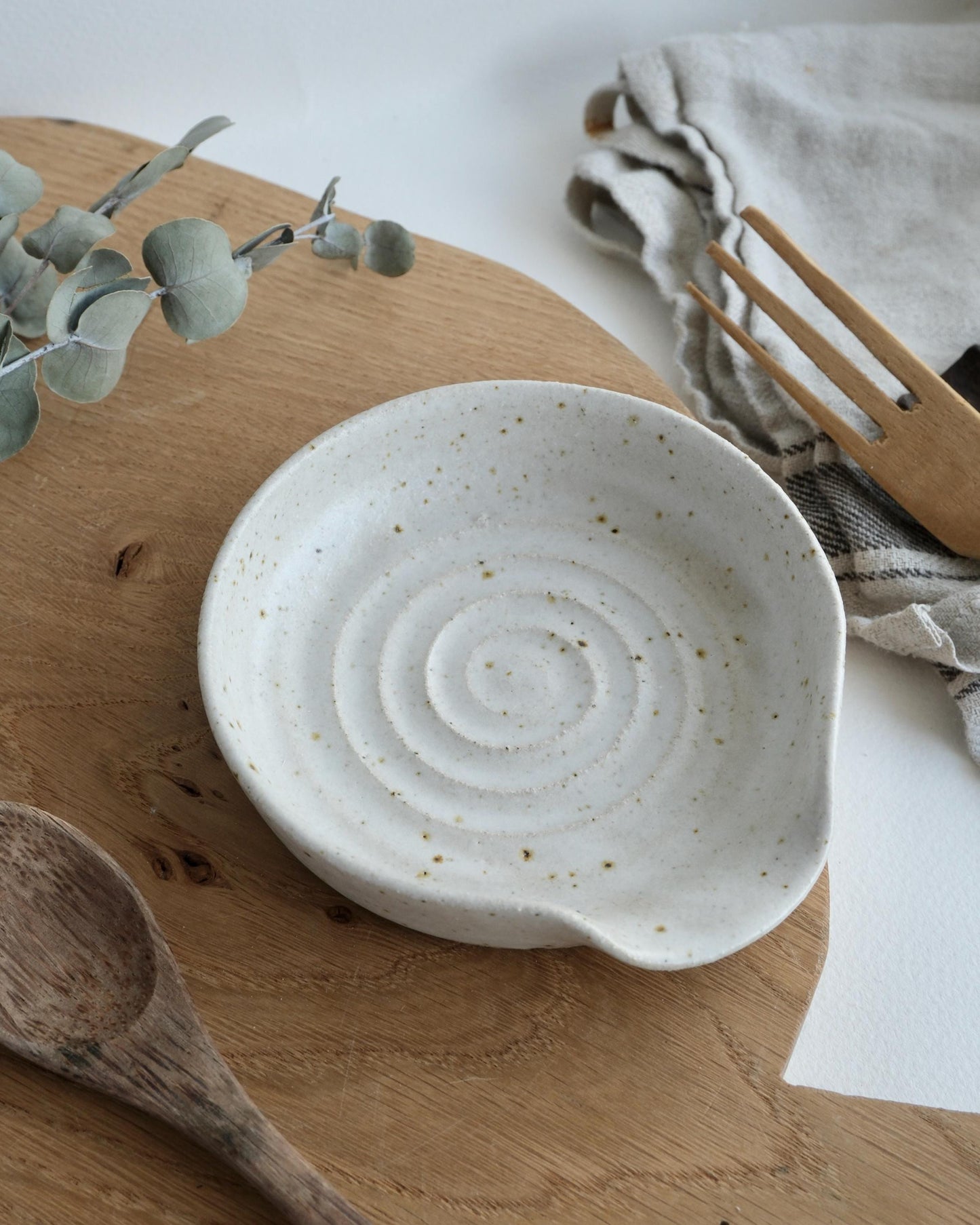 White ceramic spoon rest dish on a wooden surface with eucalyptus leaves and a wooden spoon.