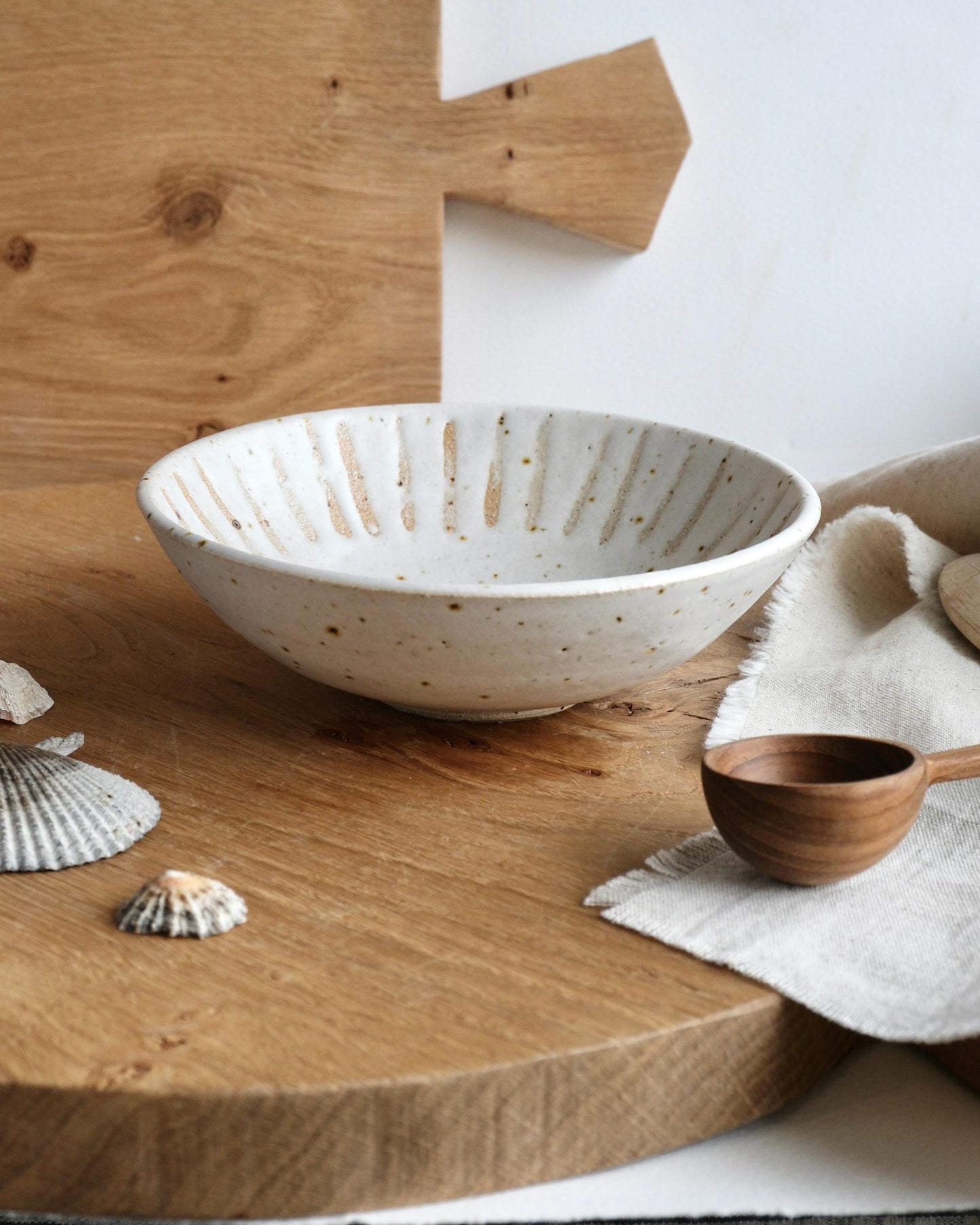 White speckled ceramic trinket bowl on a wooden surface with a wooden spoon and seashells.